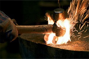 Blacksmith hammering red hot steel on a wooden surface that is catching on fire. Focus is on the hammer and glove.